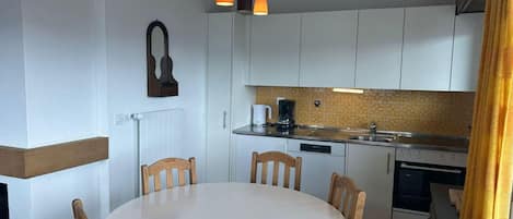 A compact kitchen with a round dining table, wooden chairs, a coffee maker, and a hanging light fixture.