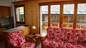 L’intérieur chaleureux d’un chalet avec un coin repas en bois, un canapé recouvert d’un tissu à motifs rouges et blancs, et de grandes fenêtres offrant une vue sur un toit en bois.