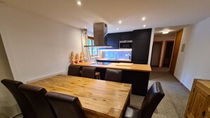 Une cuisine moderne dotée d’un îlot central, de meubles de rangement aux couleurs sombres et d’un coin repas avec une table et des chaises en bois.