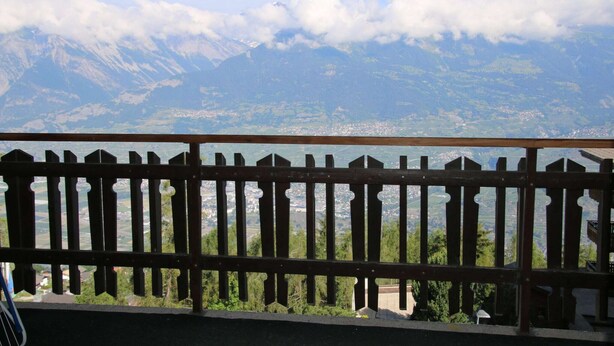 Un balcon offrant une vue sur un paysage montagneux, avec des sommets enneigés et une ville en contrebas.