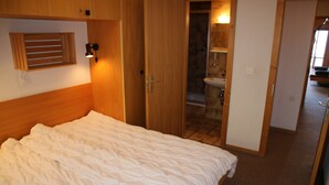 Une chambre à coucher avec une tête de lit en bois, une couvre-lit blanche matelassée et une porte en bois donnant sur une salle de bain carrelée, équipée d’un lavabo.