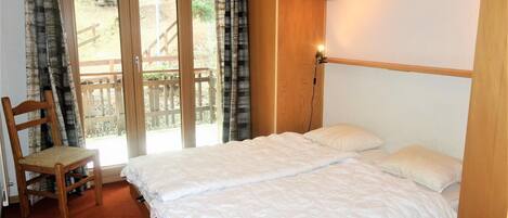 Une chambre avec un lit, une chaise et une vue sur une terrasse en bois, visible à travers une porte vitrée.