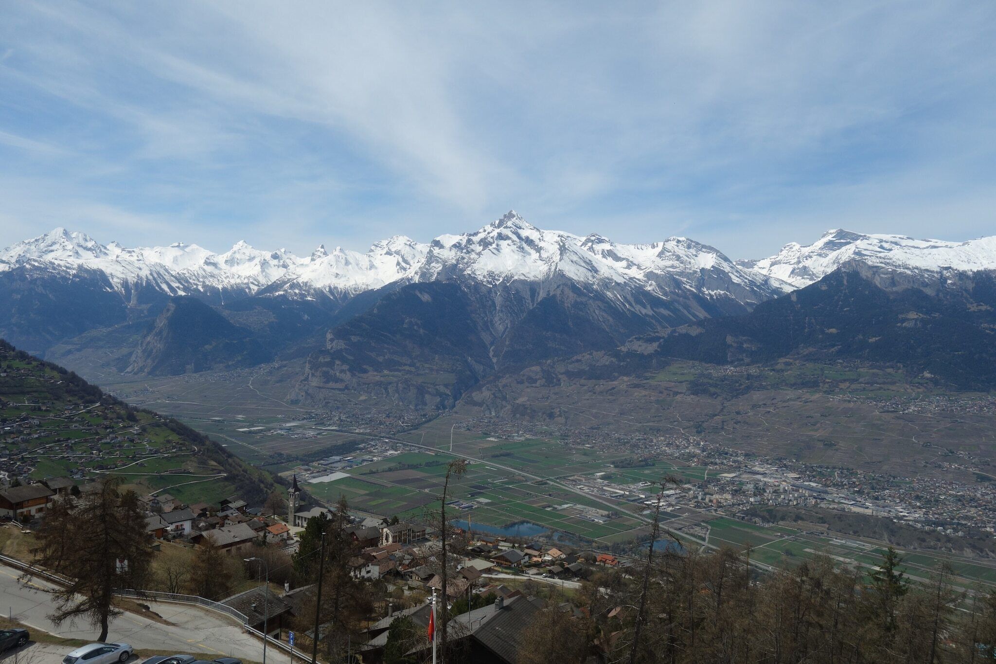 Eine gebirgige Landschaft mit schneebedeckten Gipfeln, einem Tal mit Feldern und einer kleinen Stadt mit Gebäuden und Straßen.