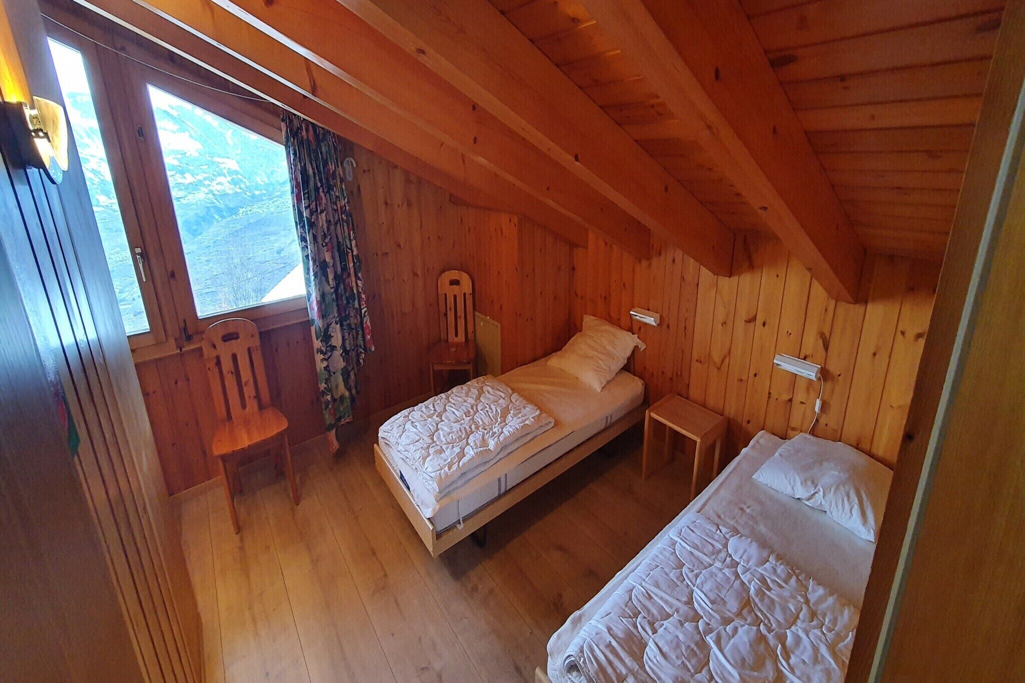 A wooden cabin room with two beds, a chair, a small table, and a window with curtains.