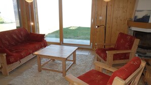Habitació acollidora amb un sofà de fusta, un taulell de fusta i dues cadires de fusta amb coixins vermells. Hi ha una finestra gran que donava a una zona verda.
