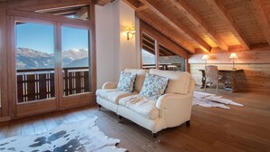 A cozy room with a wooden ceiling, a white sofa with patterned cushions, a wooden desk with a chair, and a large window offering a mountain view. - Chalet in Veysonnaz with hot tub (Veysonnaz)