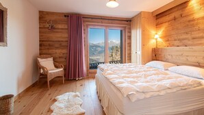 A bedroom with a wooden floor, a bed with white bedding, a wicker chair, a window with a view of mountains, and a lamp on the wall. - Chalet in Veysonnaz with hot tub (Veysonnaz)