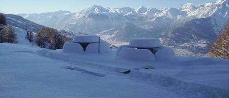 Un paysage enneigé, avec des montagnes en arrière-plan et deux constructions recouvertes de neige au premier plan.