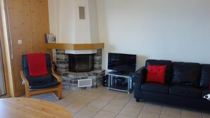 A living room with a stone fireplace, a black leather sofa with red cushions, a wooden chair with a red throw, and a television on a stand. - Spacious apartment with fireplace (Veysonnaz)