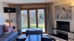 A modern living room with a sofa, a small table, a TV, a fireplace, and a large window with curtains. - Apartment in Veysonnaz with Private Balcony (Veysonnaz)