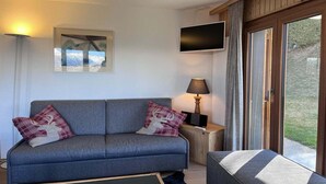 A modern living room with a grey sofa, two decorative pillows, a mounted flat-screen TV, a side table with a lamp, and a large window offering a view of the outdoors. - Apartment in Veysonnaz with Private Balcony (Veysonnaz)