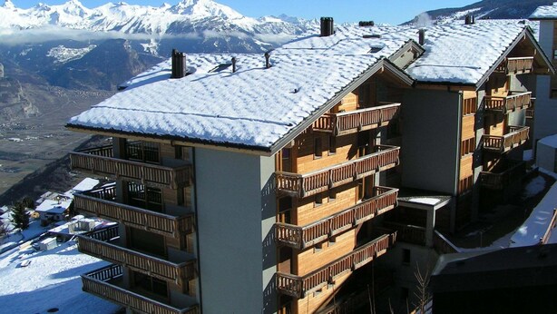 Ein mehrstöckiges Hotel mit Holzbalkonen und einem mit Schnee bedeckten Dach, vor einer Kulisse schneebedeckter Berge.