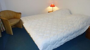 A bed with a white quilt, a wooden nightstand with a red lamp, and a brown leather chair. - Centrally-located apartment in Veysonnaz (Valais)