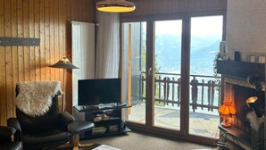 A cozy living room with a fireplace, a television, and a view of the mountains through large sliding glass doors. - Centrally-located apartment in Veysonnaz (Valais)