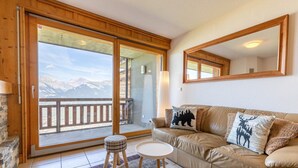En stue med en sofa, et rundt bord, en skammel og en stor skydedør, der fører ud til en balkon med udsigt til et bjerglandskab.