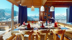 A wooden dining room with a table set for four, large windows offering a mountain view, and wooden ceiling beams.