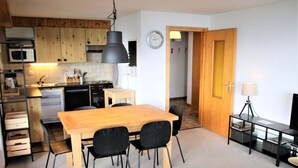 A compact kitchen with wooden cabinets, a wooden dining table, and black chairs. A clock and a lamp are visible on the walls. - Apartment in Veysonnaz near Cable Car (Veysonnaz)