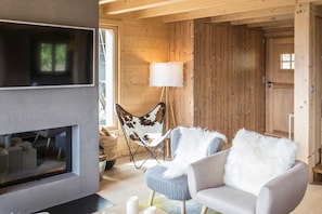 Un acogedor interior de cabaña con chimenea, un ventanal grande, un sillón con una manta de piel y una lámpara.