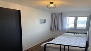 Quarto pequeno com uma cama de solteiro, piso de madeira, estrutura de cama de metal preto, colchão branco, luminária no teto, janela com cortinas e um pequeno quadro na parede.