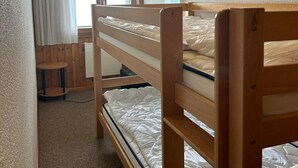 Uma cama beliche com estrutura de madeira, uma mesinha redonda e uma janela com cortinas.