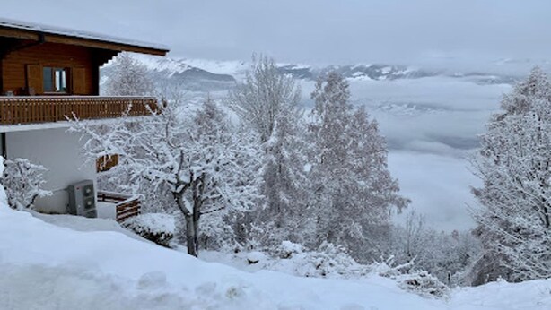Una cabaña de madera con balcón, rodeada de árboles cubiertos de nieve y montañas al fondo.