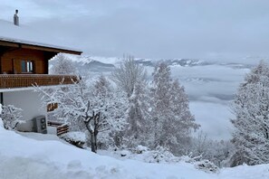 Una cabaña de madera con balcón, rodeada de árboles cubiertos de nieve y montañas al fondo.