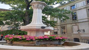 Une fontaine classique, avec une colonne et un parterre de fleurs au centre, entourée d’une zone pavée et de bâtiments en arrière-plan.