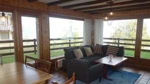 Habitación con piso de madera, sofá, mesa y una silla. Amplias ventanas ofrecen vistas al exterior.