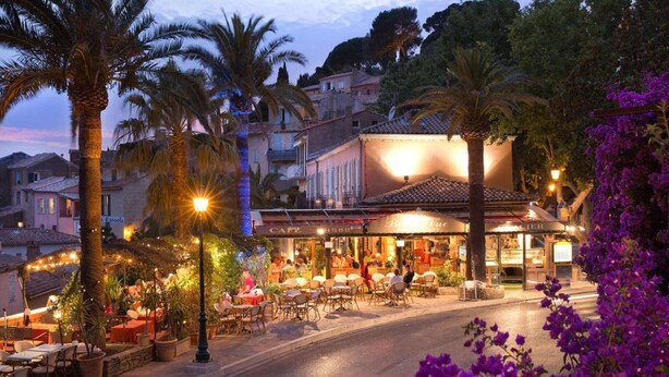 An outdoor café with tables and chairs, surrounded by palm trees and buildings.