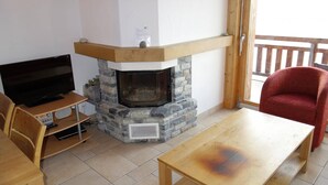 Uma sala de estar com lareira de pedra, televisão de tela plana, móveis de madeira e uma varanda com uma cadeira vermelha.