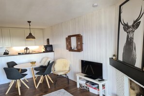 Una cocina moderna con armarios blancos, un comedor con una mesa redonda y sillas, un televisor sobre un mueble bajo y un cuadro de un ciervo en la pared.