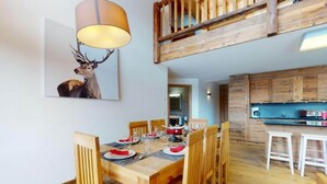 A dining area with a wooden table set for six, a painting of a deer on the wall, and a kitchen in the background.