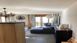 A modern living room with a sofa, a wooden cabinet, a dining table, and a large window with curtains. - Apartment in Swiss Alps near Cable Car (Veysonnaz)