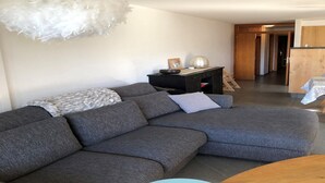 A modern living room with a grey sectional sofa, a white feathered light fixture, and a small wooden table. - Apartment in Swiss Alps near Cable Car (Veysonnaz)