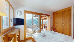 A bedroom with a bed, wooden desk, TV, and a view of mountains through a sliding door. - Lush chalet in Heremence with sauna (Hérémence)
