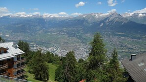 Vista de una montaña con un pueblo abajo, un edificio con balcón y picos nevados en la distancia.