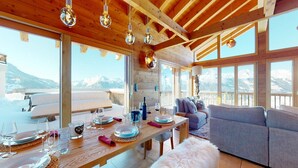 Un interior de madera con una mesa de comedor lista para una comida, un sofá y ventanas grandes con vistas a la montaña.