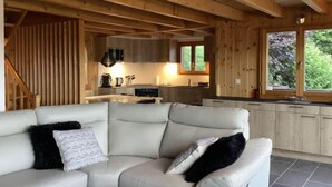 Interior moderno de una cabaña de troncos con sala de estar que incluye un sofá de cuero blanco y cocina con armarios de madera y un ventanal grande.