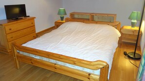 Una cama de madera con colchón blanco, dos mesitas de noche con lámparas, un aparador de madera y un televisor sobre él.