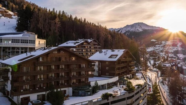 Un centro de esquí con edificios de varios pisos, techos cubiertos de nieve y montañas al fondo.