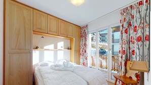 Ett sovrum med en säng, träskåp, ett skrivbord, en stol och utsikt över en balkong där en bil står parkerad.