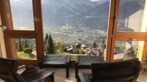 A room with large windows offering a view of a mountainous landscape and a town below. There are three black chairs in the room. - Modern apartment with balcony, ski access (Veysonnaz)