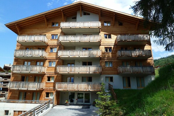 A multi-story wooden building with balconies, a central entrance, and a surrounding grassy area.