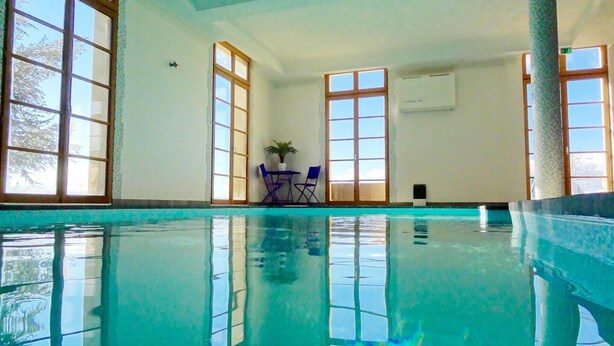 An indoor swimming pool with clear glass walls, a table with chairs, and a potted plant. - Château Comfort in Provence (Ferrassières)