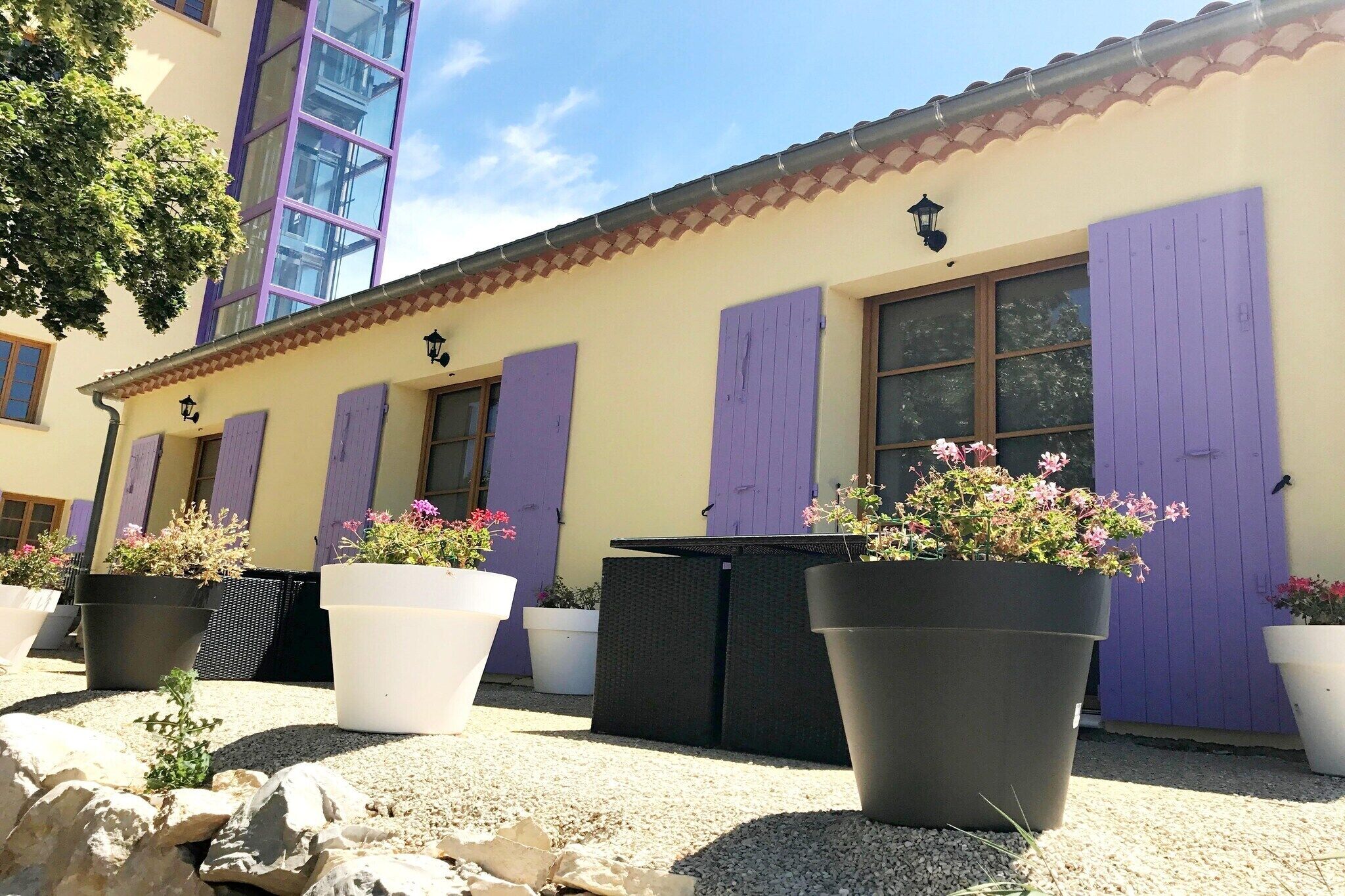 Ein Gebäude mit violetten Fensterläden, großen Kübelpflanzen und einem Ziegeldach.