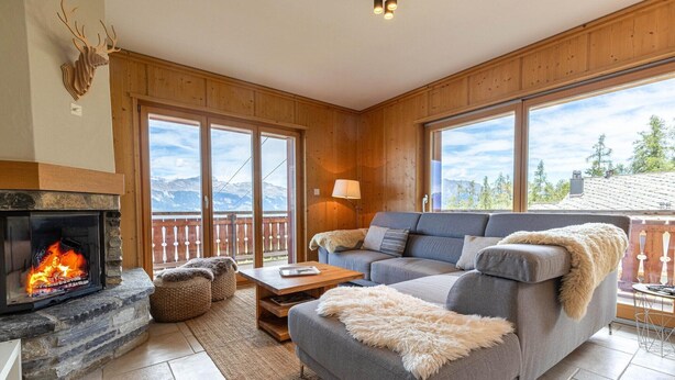 Una sala de estar acogedora con chimenea de piedra, un sofá gris y vistas a montañas a través de amplias ventanas.