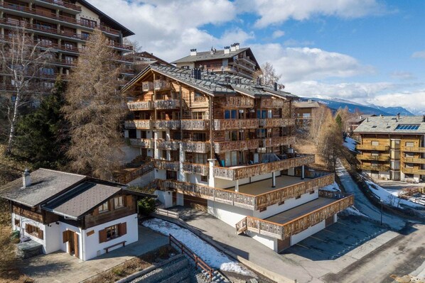 A multi-story wooden building with balconies, a small annex, and a view of snow-covered mountains.