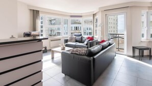 A modern living room with a black sofa, a coffee table, and a view of a cityscape through large windows. - Apartment in Ostend near Sandy Beaches (Oostende)