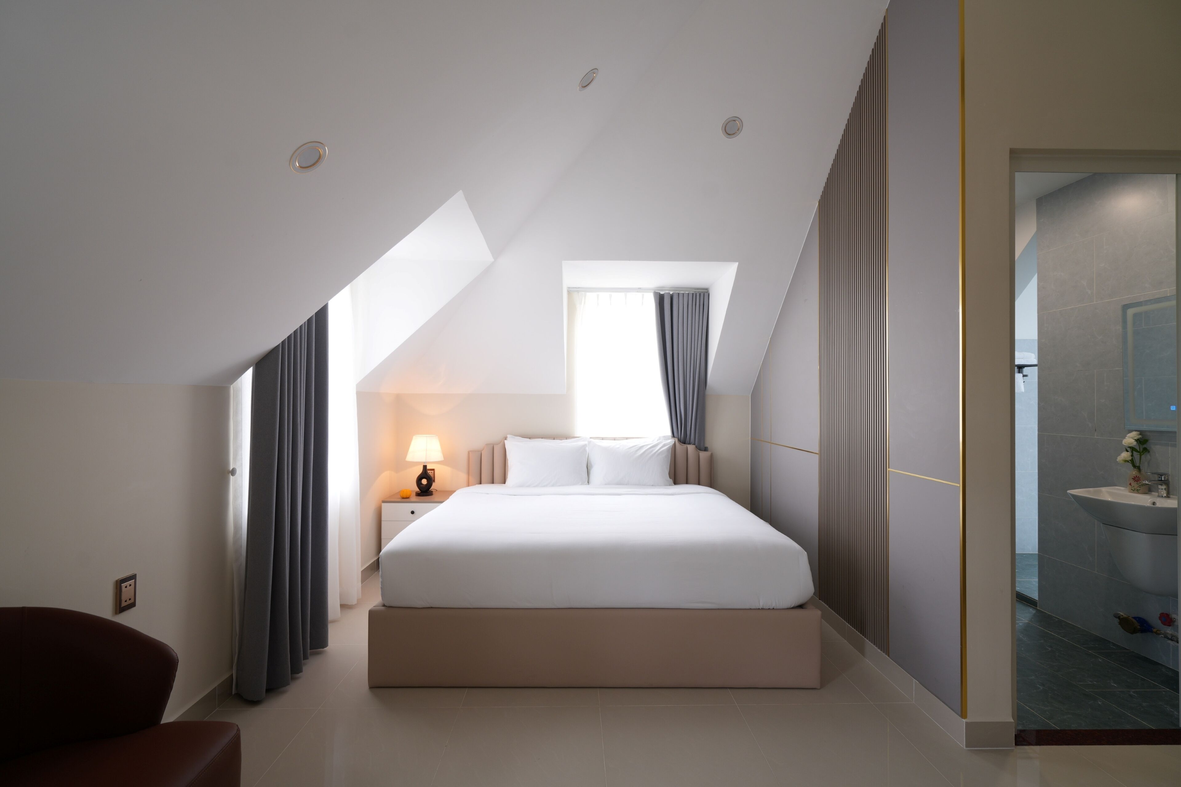 A modern hotel room with a large bed, a bedside table with a lamp, a window with curtains, and a view of the bathroom.