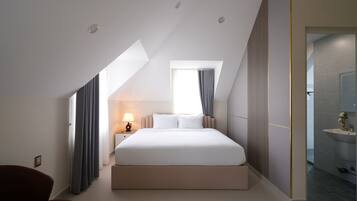 A modern hotel room with a large bed, a bedside table with a lamp, a window with curtains, and a view of the bathroom.
